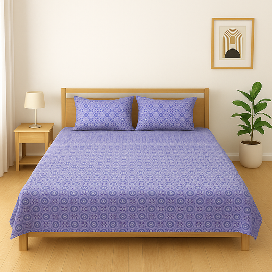 Bed with purple patterned bedding in a room with a lamp, plant, and wall art.